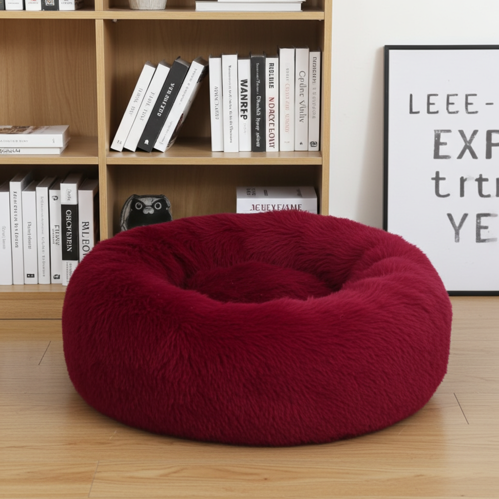 Red donut-shaped pet bed on a wooden floor with a bookshelf in the background